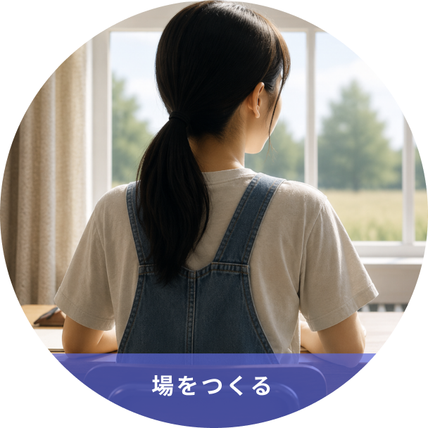 Back view of a person with dark hair in denim overalls and a light shirt, seated at a table by a window, looking out at a sunny field.