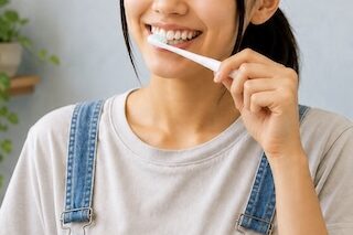 Young woman wearing a gray t-shirt and denim overalls brushing her teeth with a white toothbrush.